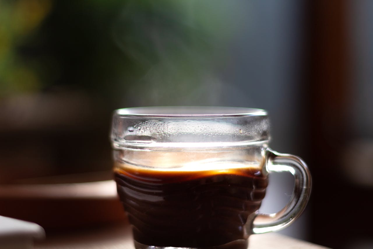 Delicious aromatic black coffee with steam in creative design glass cup with shiny surface on table for breakfast on blurred background